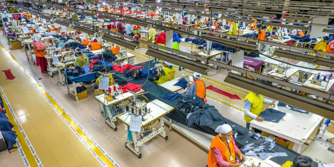 Workers in a cloth manufacturing factory working at sewing machines with clothes and threads surrounding them